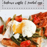 A hashbrown waffle sits on a white plate covered by asparagus, tomatoes and a poached egg being split by a fork