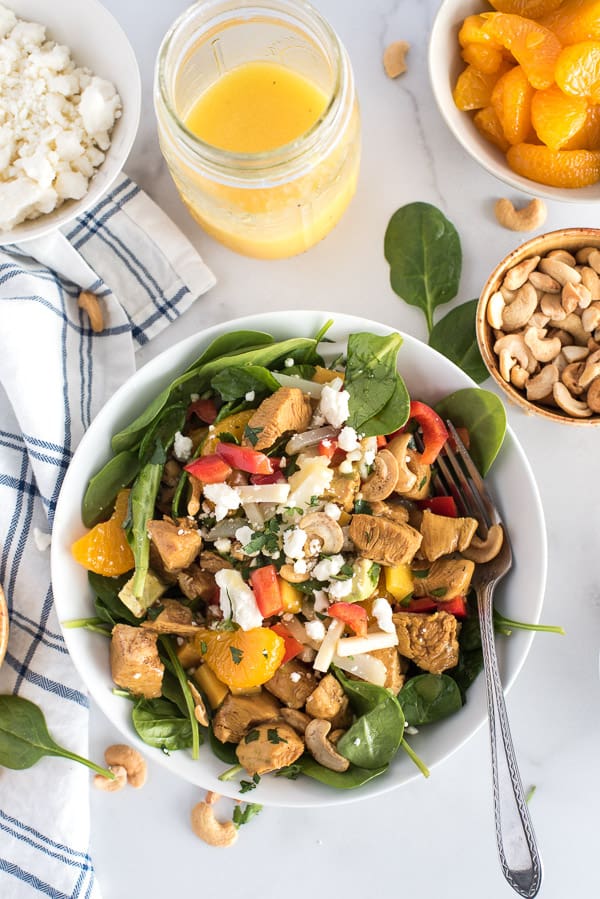 bowl of spinach salad with chicken, bell peppers, mandarin oraanges, and cheese with a bowl of cashews