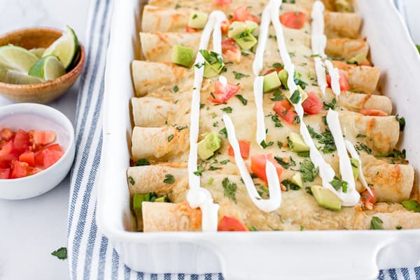 White casserole dish of chicken enchiladas with tomatoes, avocados, and sour cream