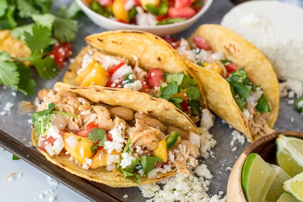 three tacos with chicken and peach salsa next to a bowl of beans and cilantro