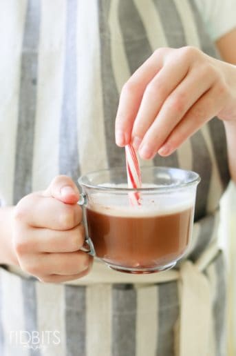 cup of hot chocolate with whipped cream in a glass cup being stirred with a candy cane