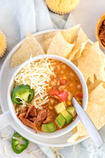 chicken chili in a white bowl with avocado, bacon, cheese, and jalapenos and a side of chips