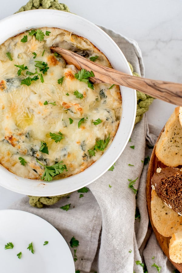 spinach artichoke dip in a white bowl being scooped with a wooden spoon