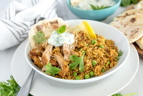 Chicken and rice in a white bowl with naan, peas, and lemon.